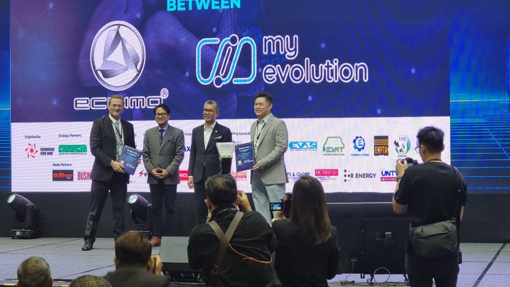 Four men stand on a stage during the signing ceremony of a Memorandum of Understanding (MOU) between Eclimo and My Evolution. The backdrop features the logos of both companies along with several sponsor logos. Two of the men hold signed documents, while photographers capture the moment. The event appears formal, with participants dressed in business attire.